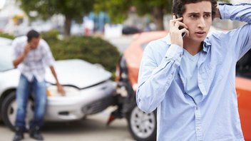 Un homme au téléphone semble avoir eu un accident avec sa voiture, qui se trouve en arrière plan.