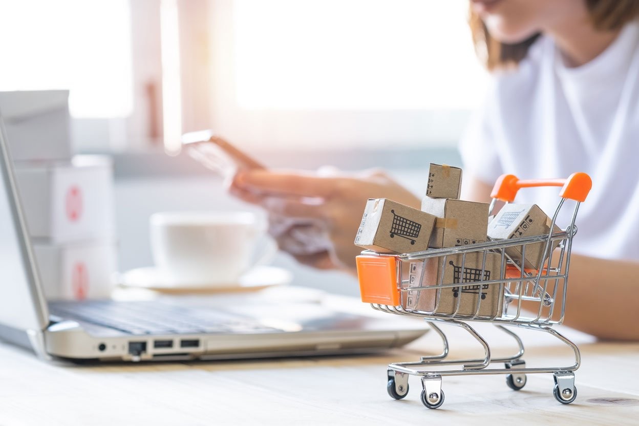 Eine Frau kauft online ein; vor ihr steht ein kleiner Einkaufswagen mit Paketen