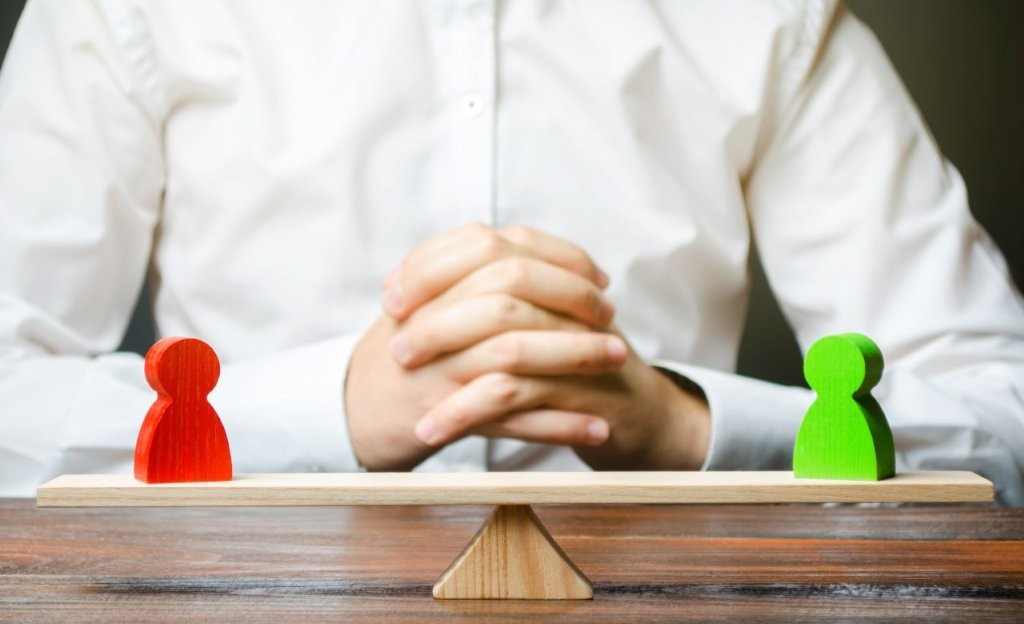 Mann faltet die Hände, vor ihm stehen eine grüne und eine rote Figur auf einer Holzwaage im Gleichgewicht