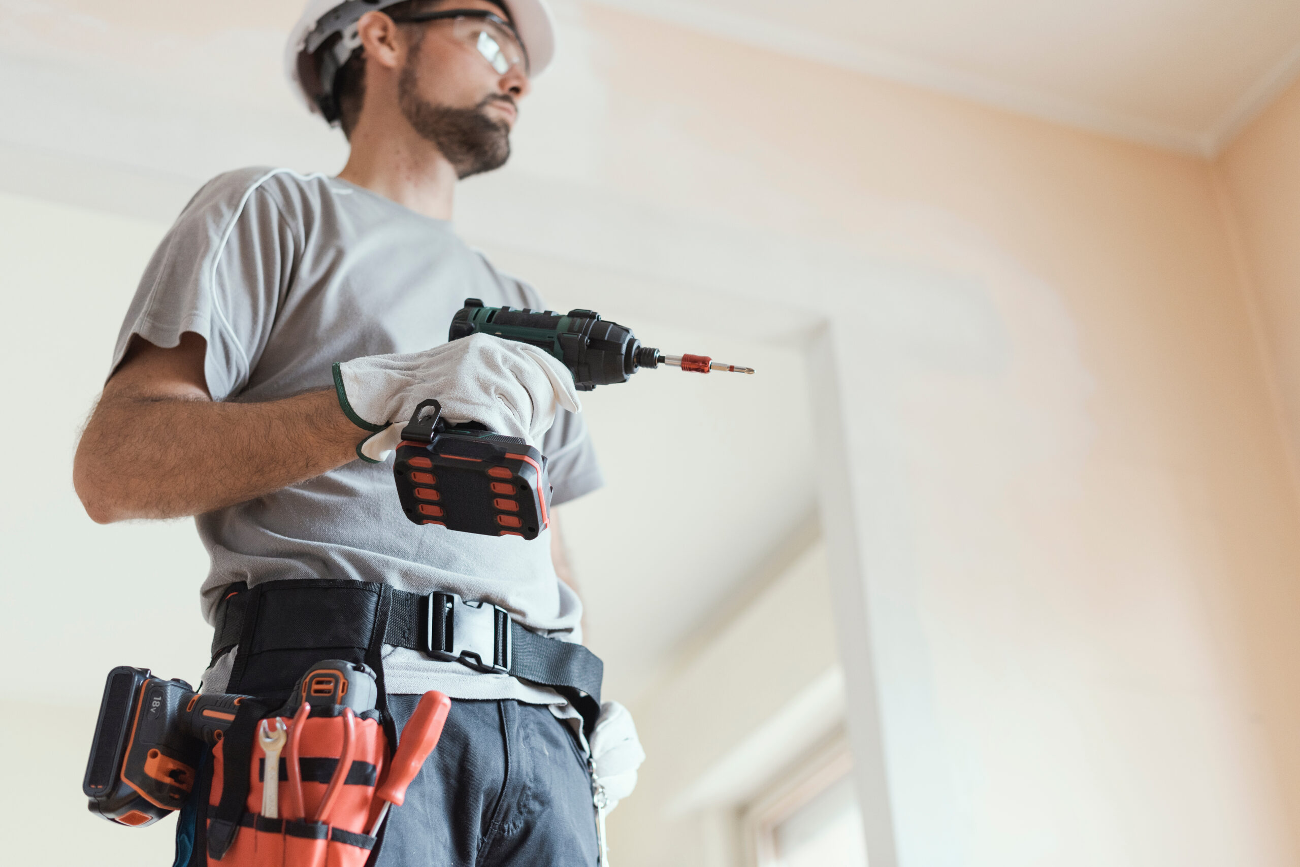 Un ouvrier avec un casque sur la tête et une perceuse dans la main réalise des travaux à l'intérieur d'un logement