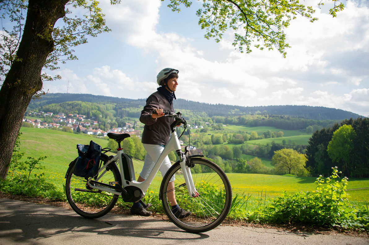 Eine Frau mit einem Fahrradhelm schiebt ein weißes Pedelec auf einem Weg.