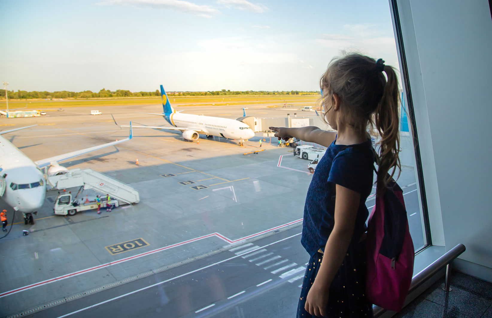 Une jeune fille est dans le hall de départ d'un aéroport et pointe du doigt un avion sur le tarmac