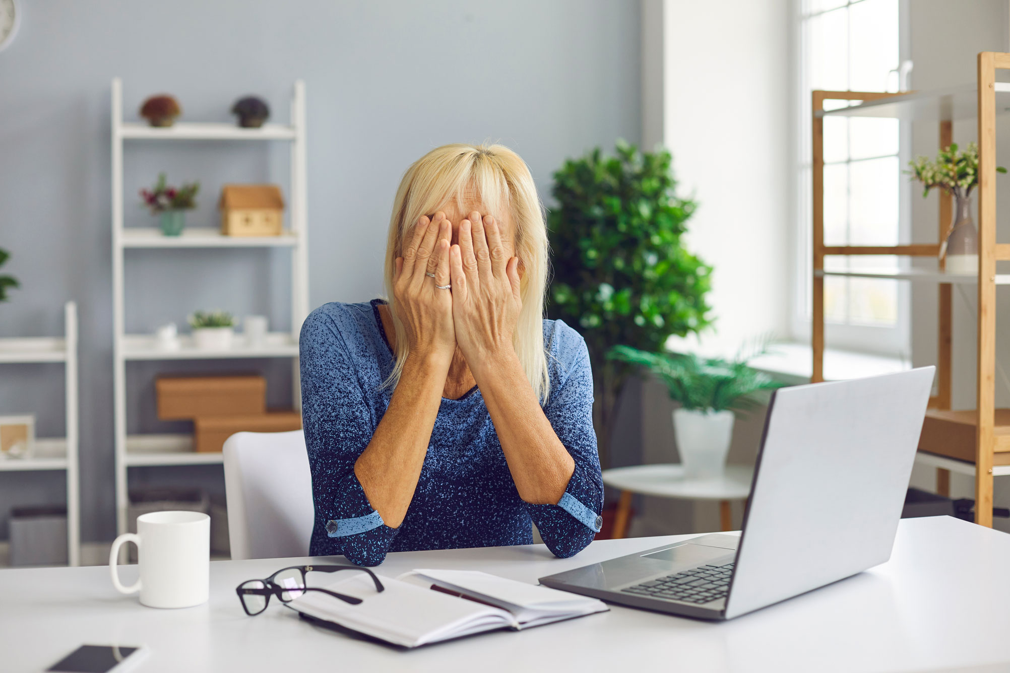 Une femme, assise devant une table avec un ordinateur cache son visage avec ses mains, illustration d'une personne victime d'une arnaque au crédit sur internet.