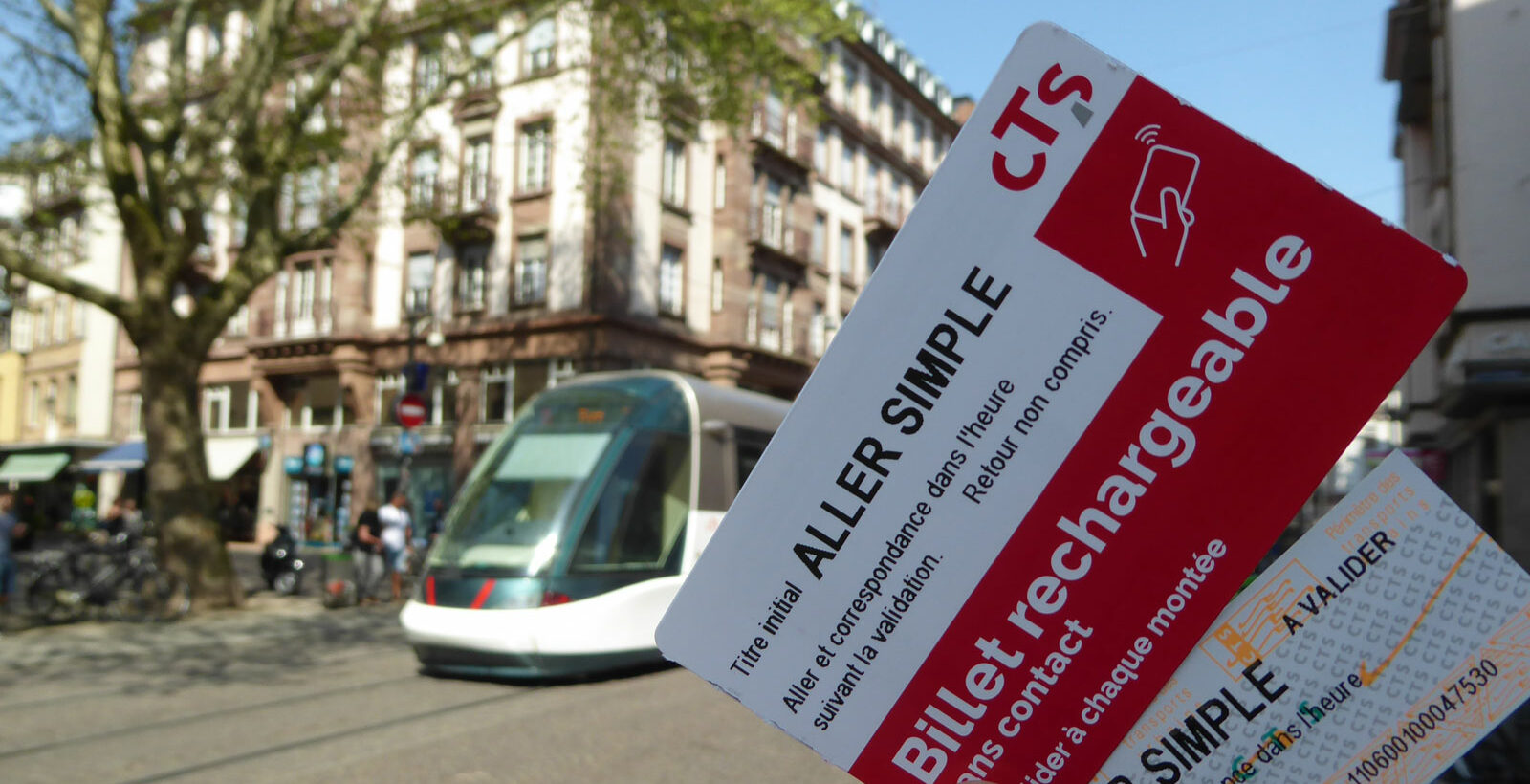 Billet simple et un billet rechargeable de la CTS tenus dans un main au centre de Strasbourg pendant qu'un tram passe en arrière-plan
