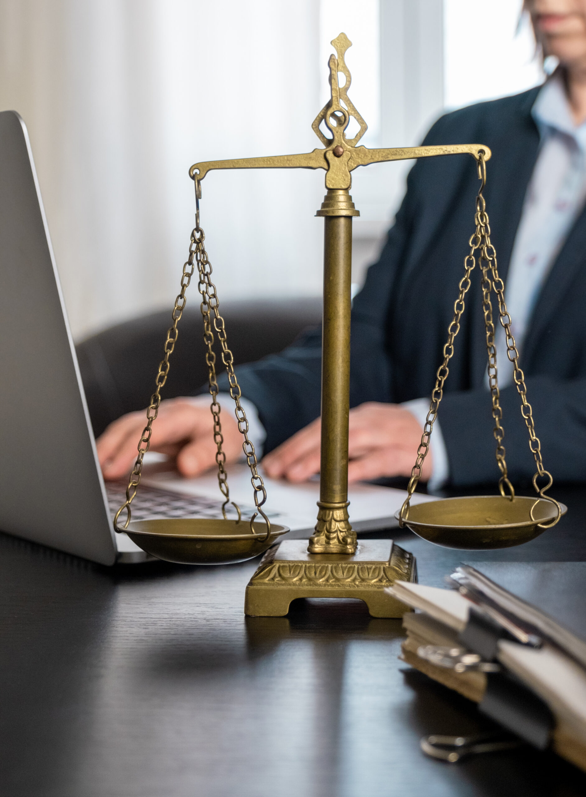 Eine Person arbeitet am Laptop, auf dem Schreibtisch vor ihr Unterlagen und eine Waage als Symbol der Justiz.