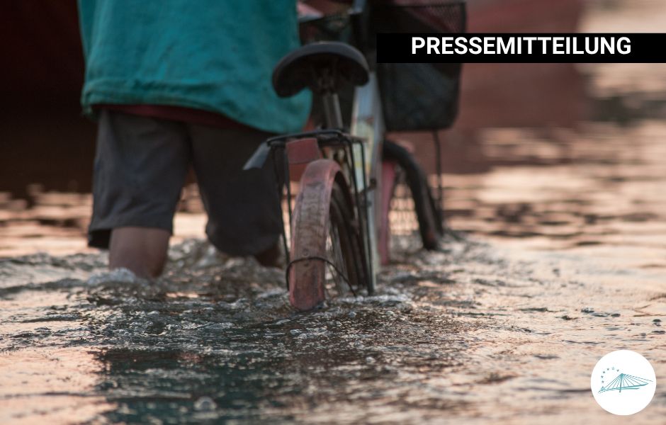 Eine Person in kurzer Hose und Regenjacke schiebt ein Fahrrad durch kniehohes Wasser nach einer Hochwasser-Episode.