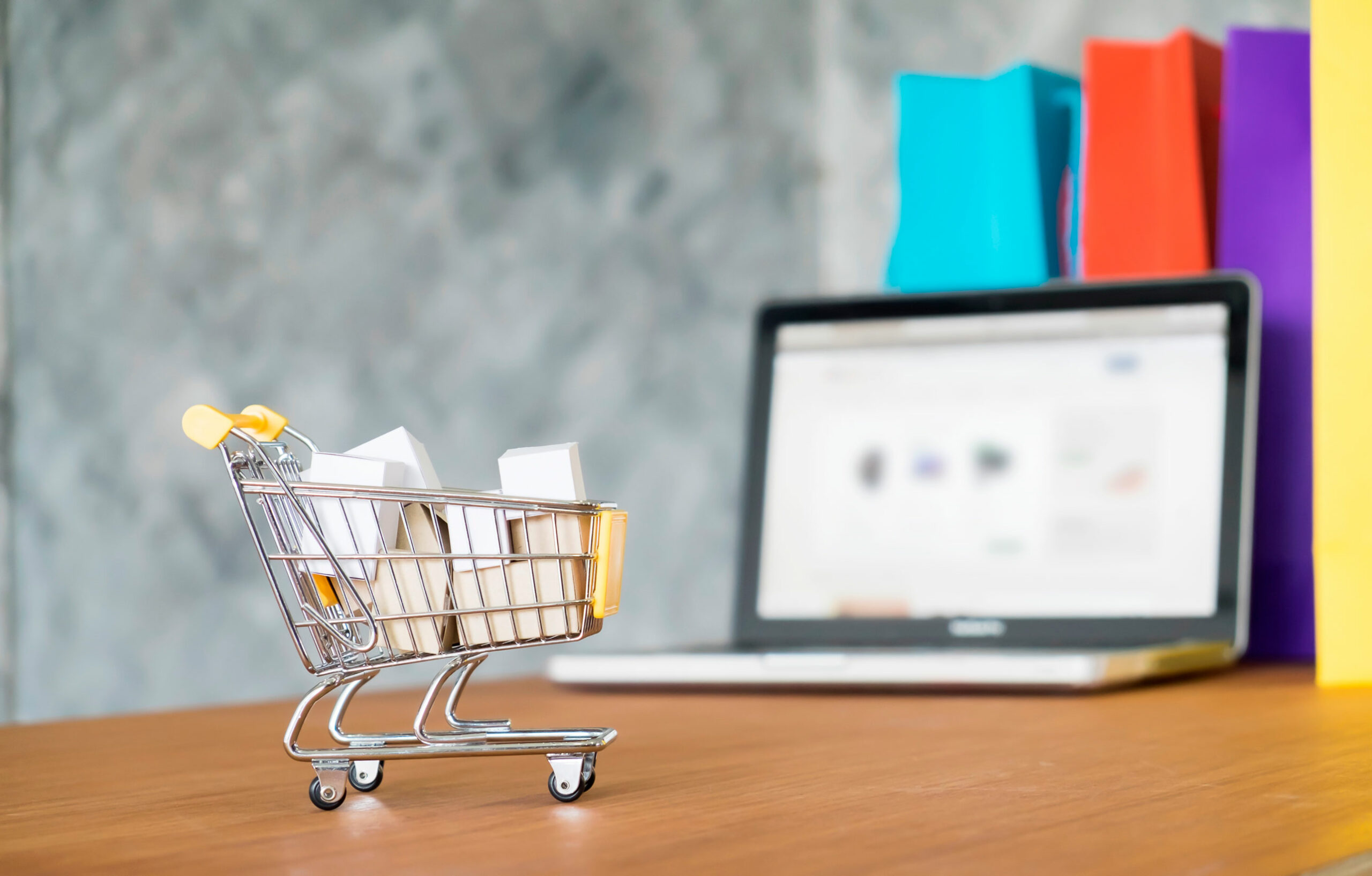 Illustration d'un achat avec un caddie miniature rempli de colis et posé sur une table devant un ordinateur