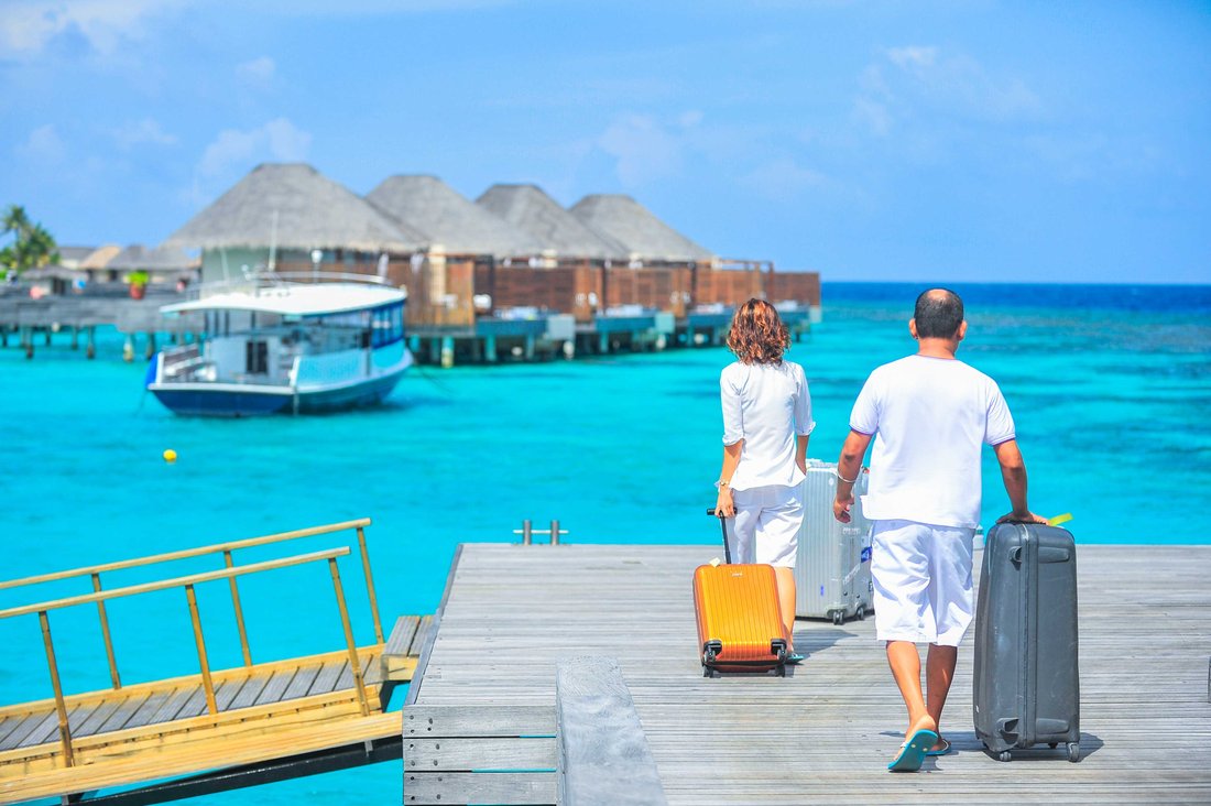 Un couple qui marche valises à la main sur une passerelle au dessus d'une eau turquoise. On voir en arrière plan des bungalow en bois et en paille.